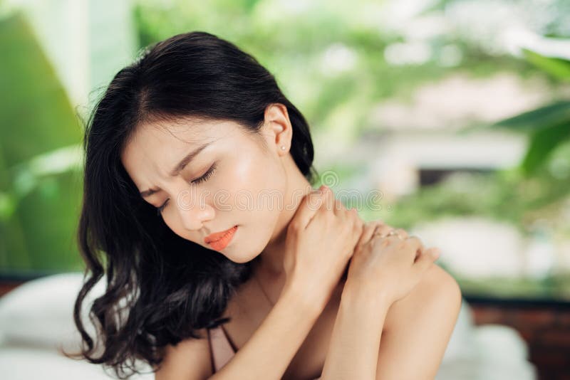 Ungt asiatiskt kvinnalidande från hals smärtar, medan sitta på säng royaltyfri foto Ungt asiatiskt kvinnalidande från hals smärtar, medan sitta på säng royaltyfri foto