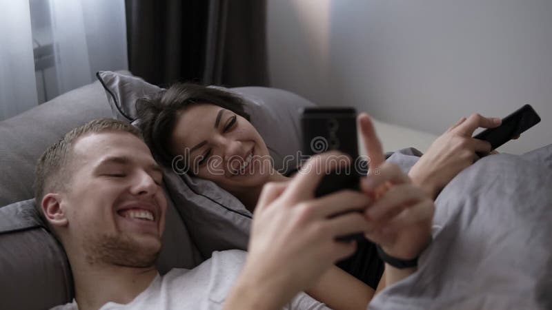 Ungt gift par som använder deras mobiltelefon, medan ligga i säng och le till varandra Lycklig livsstil, par, säng