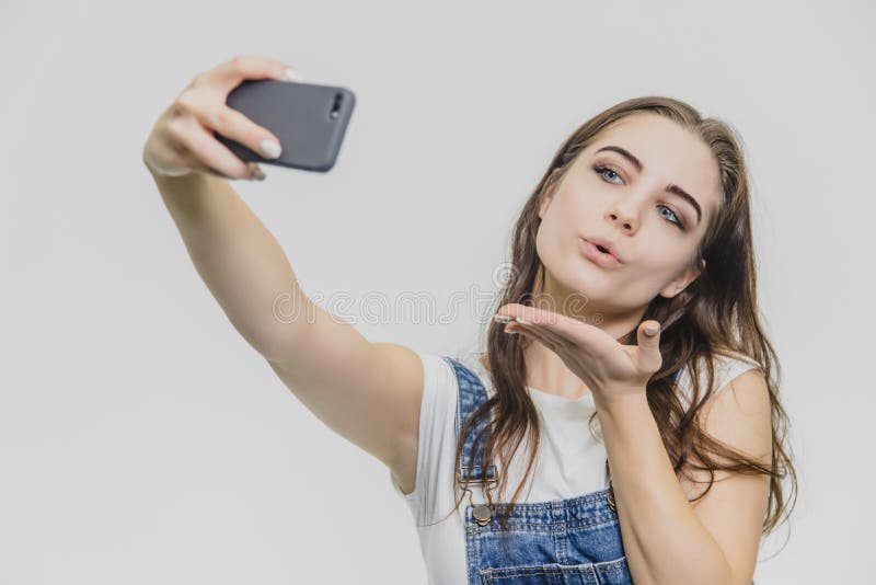 Ungt n?tt flickaanseende p? en vit bakgrund Iklädda blåa grov bomullstvilloveraller för a och en vit T-tröja När rymma arkivbild Ungt n?tt flickaanseende p? en vit bakgrund Iklädda blåa grov bomullstvilloveraller för a och en vit T-tröja När rymma arkivbild