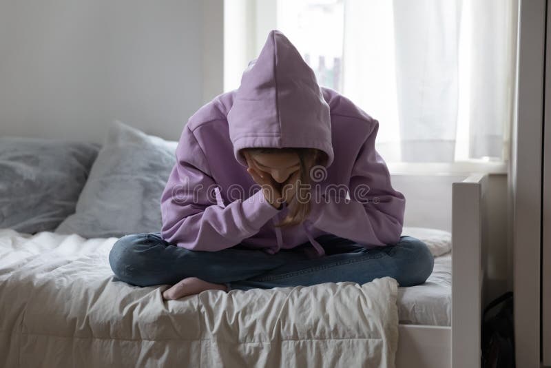 Unhappy depressed teenage girl wearing sweatshirt sitting on bed alone, feeling lonely and misunderstood, stressed sad teenager thinking about troubles, hiding face, child and psychological problem