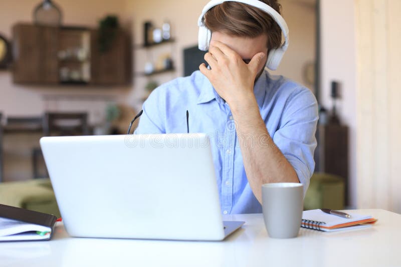 Unhappy frustrated young male holding head by hands sitting with laptop behind desk at home royalty free stock photography Unhappy frustrated young male holding head by hands sitting with laptop behind desk at home royalty free stock photography