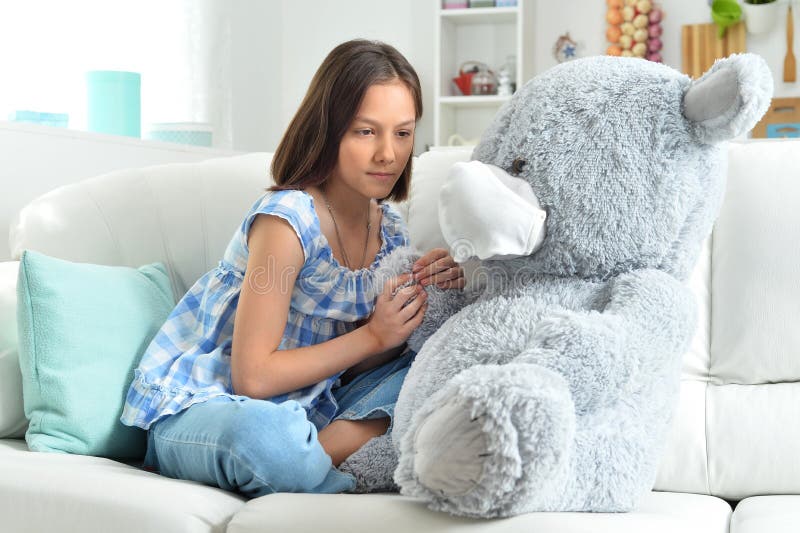 Unhappy girl with toy bear in facial mask on sofa