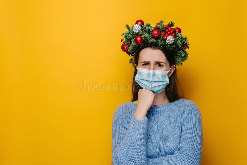 Unhappy young female wearing medical face mask, looks sad, shrugs with crossed arms, wears spruce holiday wreath around head