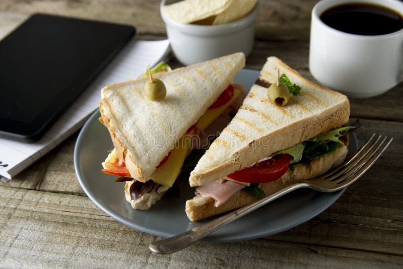 Unhealthy eatting at work place. Triangle, club sandwich with ham and vegetables. Tablet, gadget screen and notebook. Cup of royalty free stock images Unhealthy eatting at work place. Triangle, club sandwich with ham and vegetables. Tablet, gadget screen and notebook. Cup of royalty free stock images