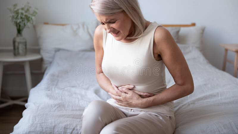 Unhealthy mature woman holding belly, feeling discomfort, health problem concept, unhappy older female sitting on bed, suffering from stomachache, food poisoning, gastritis, abdominal pain, climax. Unhealthy mature woman holding belly, feeling discomfort, health problem concept, unhappy older female sitting on bed, suffering from stomachache, food poisoning, gastritis, abdominal pain, climax