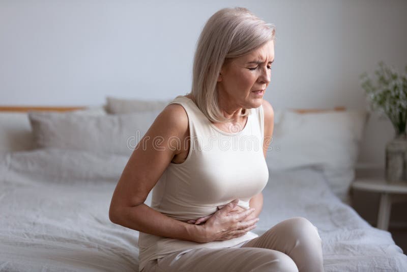 Unhealthy middle aged frowning woman sitting on bed, putting hands on belly, suffering from strong stomach ache. Unhappy older lady having painful feeling in abdomen, pancreatitis gastritis symptom. Unhealthy middle aged frowning woman sitting on bed, putting hands on belly, suffering from strong stomach ache. Unhappy older lady having painful feeling in abdomen, pancreatitis gastritis symptom.
