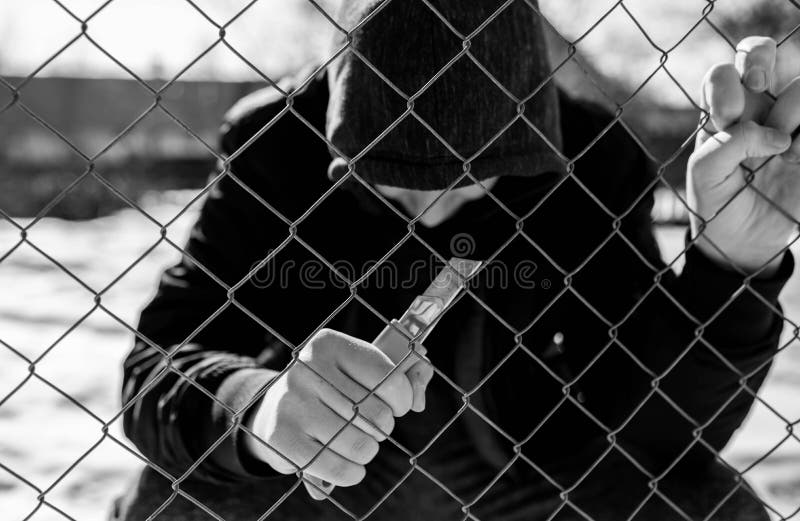 Unidentifiable teenage boy behind wired fence holding a paper knife at correctional institute, focus on the fence in black and white, conceptual image of juvenile delinquency.