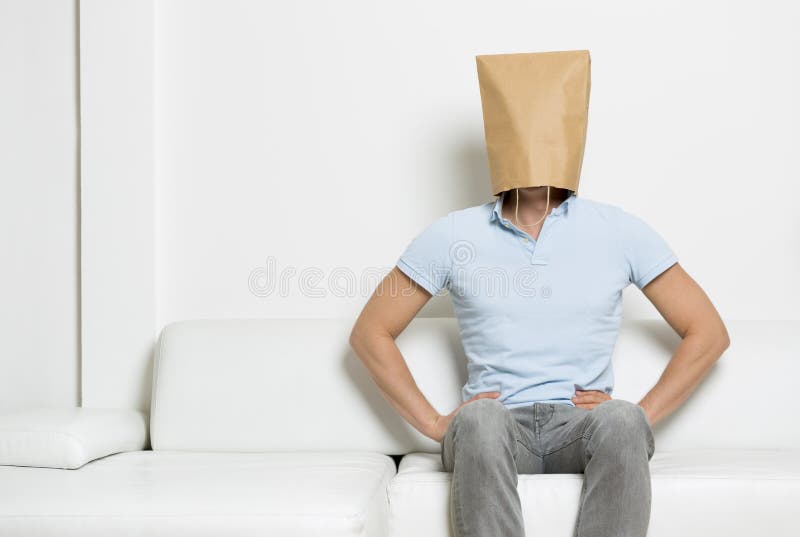 Serious anonymous man with head covered by a blank paper bag sitting on sofa with arms akimbo, empty space for text, on white background. Serious anonymous man with head covered by a blank paper bag sitting on sofa with arms akimbo, empty space for text, on white background.