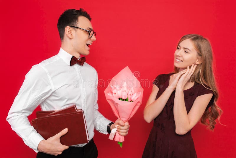 Uno studente bello, i vetri d'uso, le elasticità un regalo e un mazzo dei fiori alla sua amica contro un fondo rosso fotografie stock libere da diritti Uno studente bello, i vetri d'uso, le elasticità un regalo e un mazzo dei fiori alla sua amica contro un fondo rosso fotografie stock libere da diritti