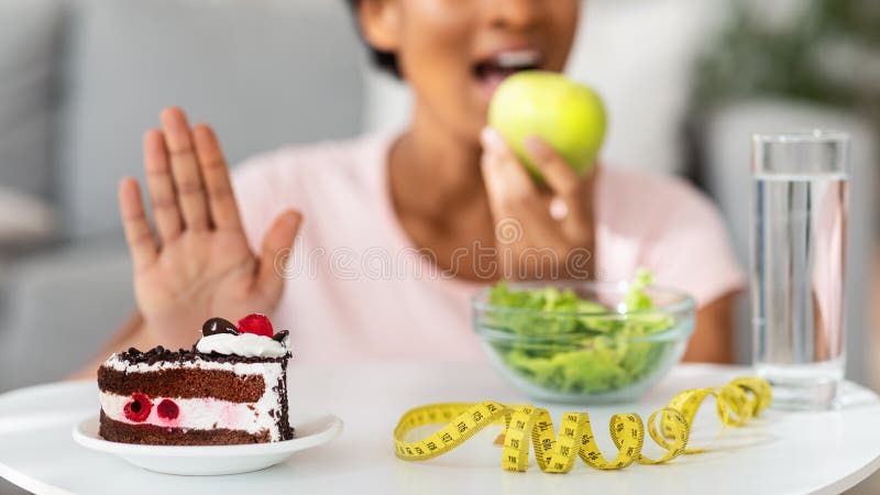 Unrecognizable black woman eating fresh apple and refusing unhealthy cake, blank space