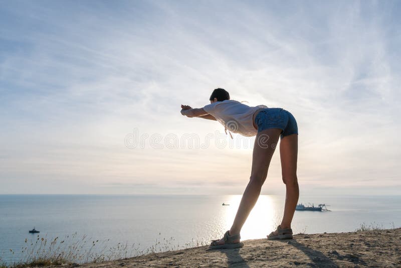 Unrecognizable kobieta w lekkiej bluzce i drelichu zwiera rozciąganie na wzgórzu z seascape Bocznym widokiem zdjęcia stock Unrecognizable kobieta w lekkiej bluzce i drelichu zwiera rozciąganie na wzgórzu z seascape Bocznym widokiem zdjęcia stock
