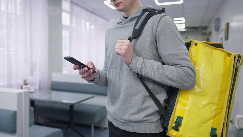 Unrecognizable male courier checking order in smartphone app standing in cafe with yellow thermal backpack. Young stock video footage