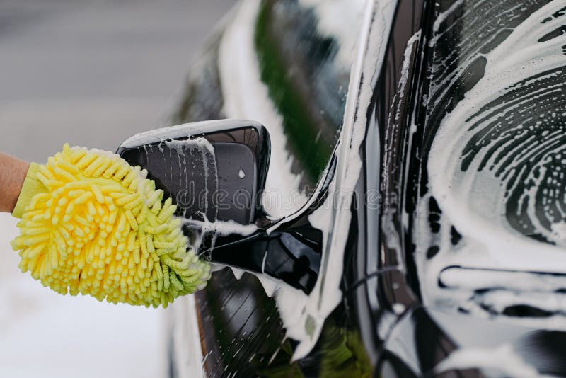 Unrecognizable man washes black car with cloth or rag mitten, uses special detergent. Cleaning dirty automobile. Carwash business concept