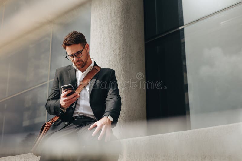 Unternehmer, der draußen Schreibenmitteilung an seinem Handy sitzt. Leitungsdraußen sitzen des geschäfts des Geschäftsmannes in Bewegung Geschäftsmann, der außerhalb eines Gebäudes betrachtet seinen Handy sitzt