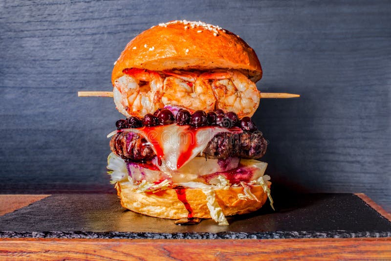 Unusual Huge burger with beef, shrimp, pineapple, blackcurrant, cheese, fresh vegetables and sauces on plate over wooden