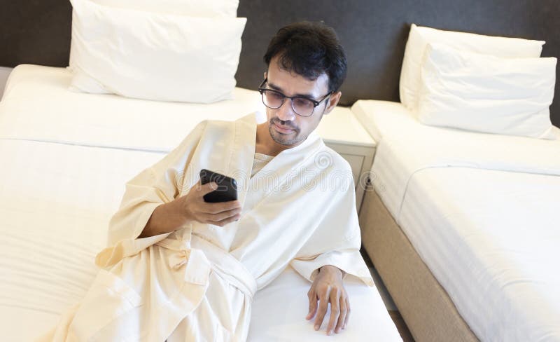 Uomo in accappatoio su letto in camera da letto e con smartphone immagine stock