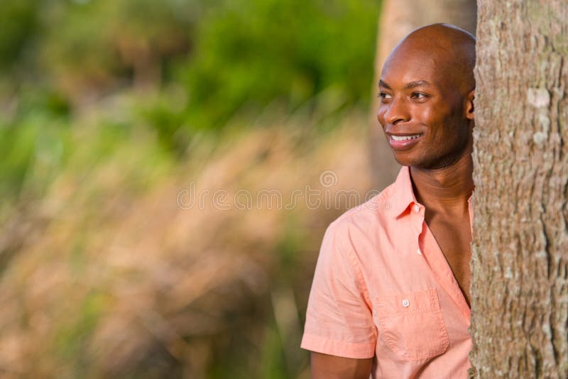 Uomo afroamericano bello del ritratto che posa da dietro un albero nel parco L'uomo è sorridente e dante un'occhiata fuori dalla fotografia stock Uomo afroamericano bello del ritratto che posa da dietro un albero nel parco L'uomo è sorridente e dante un'occhiata fuori dalla fotografia stock