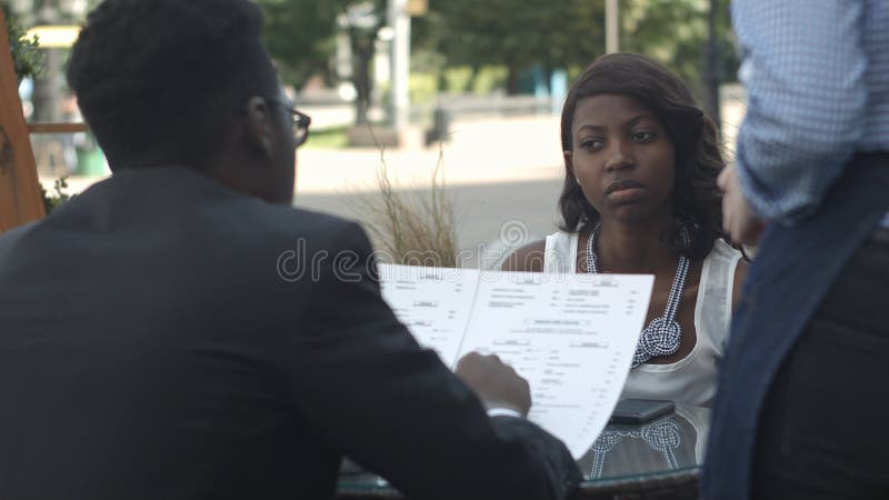 Uomo afroamericano che ordina in caffè esterno, mentre sedendosi con il suo partner femminile immagine stock Uomo afroamericano che ordina in caffè esterno, mentre sedendosi con il suo partner femminile immagine stock
