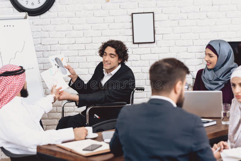 Uomo arabo disabile in sedia a rotelle che funziona nell'ufficio L'uomo sta facendo la presentazione davanti ai colleghe immagini stock Uomo arabo disabile in sedia a rotelle che funziona nell'ufficio L'uomo sta facendo la presentazione davanti ai colleghe immagini stock