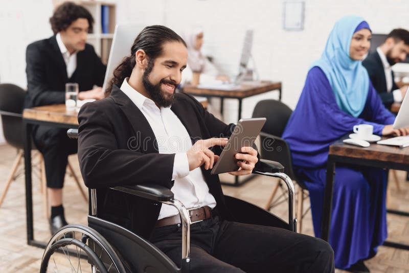 Uomo arabo disabile in sedia a rotelle che funziona nell'ufficio L'uomo sta parlando sulla compressa immagini stock Uomo arabo disabile in sedia a rotelle che funziona nell'ufficio L'uomo sta parlando sulla compressa immagini stock