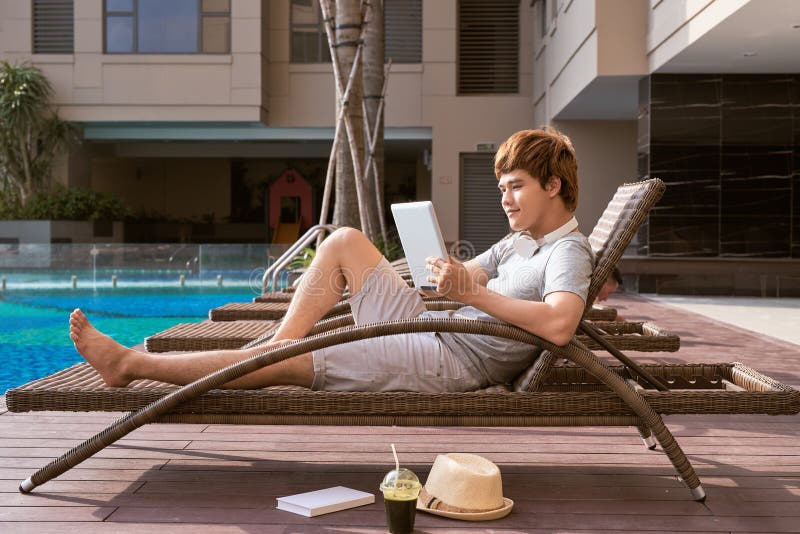 Uomo asiatico bello che si rilassa dallo stagno e che legge libro elettronico fotografia stock libera da diritti Uomo asiatico bello che si rilassa dallo stagno e che legge libro elettronico fotografia stock libera da diritti