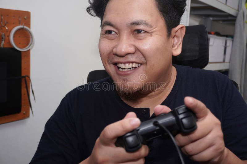 Uomo asiatico che gioca ai videogiochi con un computer felice espressione sorridente mentre regge la console immagine stock