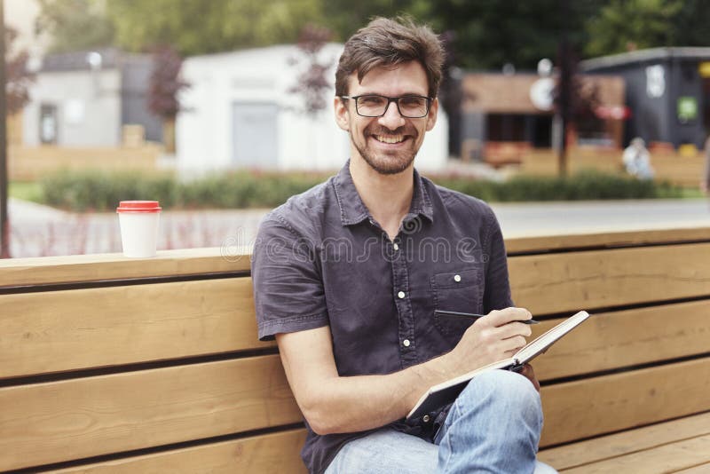 Uomo bello che fa le note in un libro che si siede fuori Lavoro solo d'uso sorridente di vetro del fronte Concetto degli studenti fotografie stock Uomo bello che fa le note in un libro che si siede fuori Lavoro solo d'uso sorridente di vetro del fronte Concetto degli studenti fotografie stock