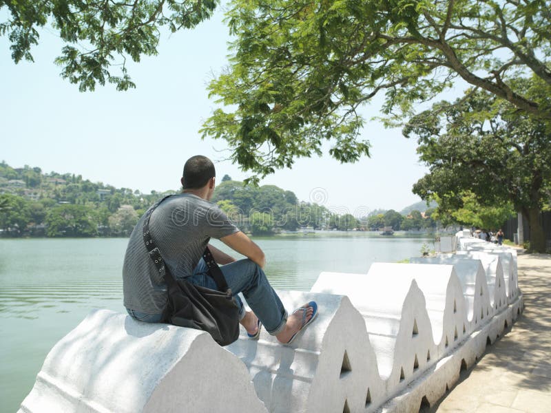 Uomo che si siede sul muro di cinta dal fiume fotografia stock Uomo che si siede sul muro di cinta dal fiume fotografia stock