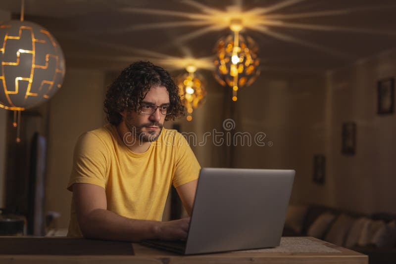 Young man sitting at kitchen counter, using laptop computer, working late, remotely from home due to social distancing pandemic prevention measures. Young man sitting at kitchen counter, using laptop computer, working late, remotely from home due to social distancing pandemic prevention measures