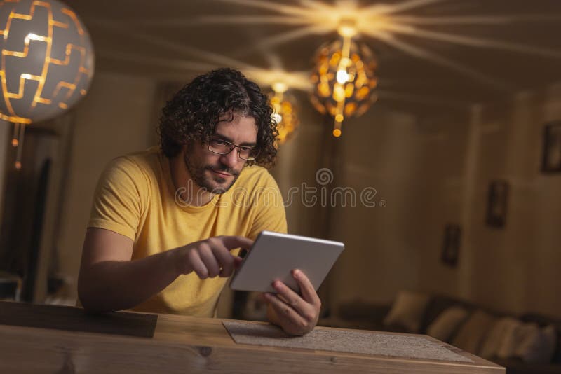 Handsome young man sitting at kitchen counter, surfing the Internet using tablet computer, relaxing at home at the end of busy work day. Handsome young man sitting at kitchen counter, surfing the Internet using tablet computer, relaxing at home at the end of busy work day