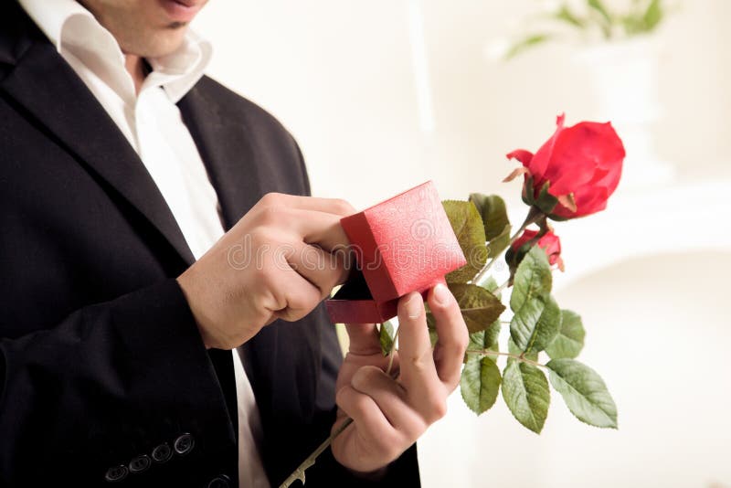 Closeup torso image of the hands of a young man about to propose holding a single long stemmed red rose and checking to make sure the ring is inside its box. Closeup torso image of the hands of a young man about to propose holding a single long stemmed red rose and checking to make sure the ring is inside its box