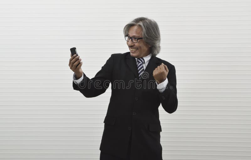 Uomo d'affari asiatico anziano emozionante felice in vestito nero ed occhiali che esaminano il suo telefono cellulare astuto e ch immagini stock libere da diritti Uomo d'affari asiatico anziano emozionante felice in vestito nero ed occhiali che esaminano il suo telefono cellulare astuto e ch immagini stock libere da diritti