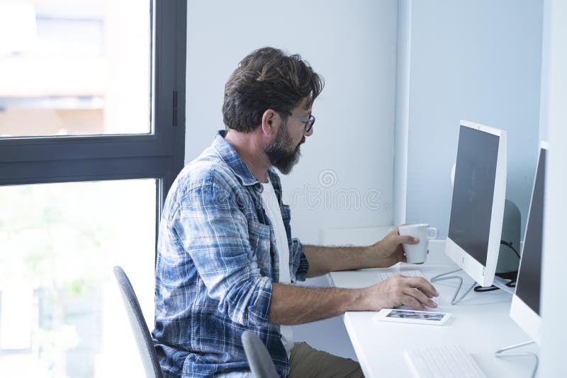 Uomo d'affari che beve caffè mentre lavora su un computer desktop. uomo maturo che usa il computer mentre sta seduto sulla sedia. uomo d'affari che usa il mouse con il cellulare sul tavolo
