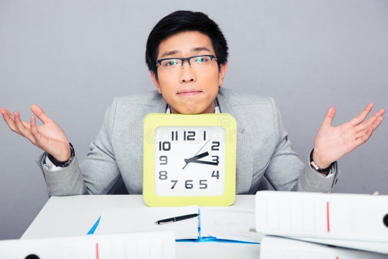 Uomo d'affari che si siede alla tavola con il grande orologio e scrollare le spalle fotografia stock libera da diritti Uomo d'affari che si siede alla tavola con il grande orologio e scrollare le spalle fotografia stock libera da diritti
