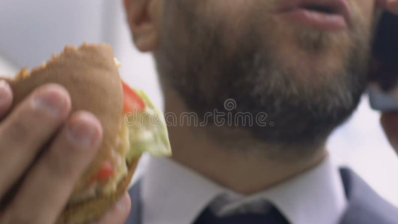 Uomo d'affari occupato che parla sul telefono e che mangia hamburger, alimenti a rapida preparazione non sani