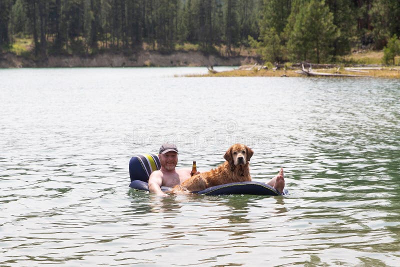 Uomo felice che beve una birra e che galleggia su un lago con il suo cane di golden retriever fotografie stock Uomo felice che beve una birra e che galleggia su un lago con il suo cane di golden retriever fotografie stock