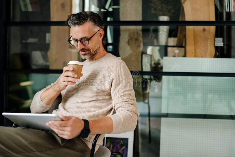 Uomo grigio adulto che beve caffè mentre lavora con il tablet