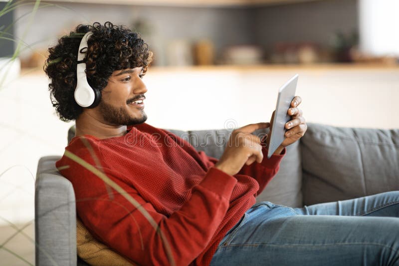 Uomo indiano sorridente con cuffie wireless poggiate sulla tavoletta digitale in casa fotografia stock libera da diritti