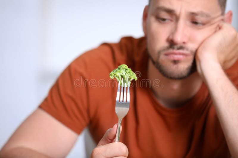 Unhappy man with broccoli on fork against light background, closeup. Unhappy man with broccoli on fork against light background, closeup