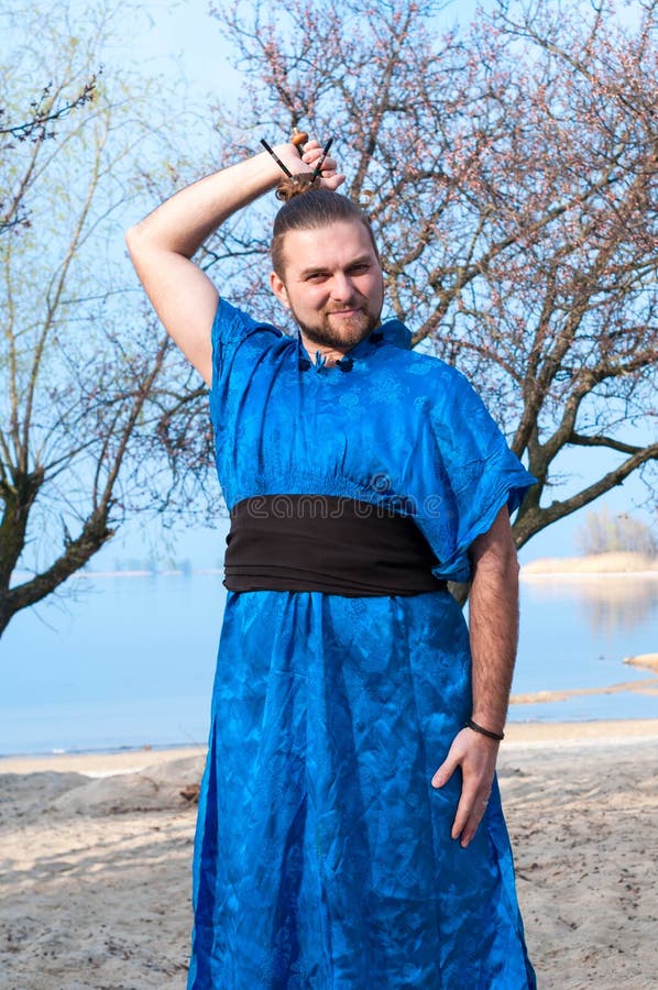 Uomo in kimono, panino e bastoni blu sulla testa che mette spada dietro la parte posteriore e che esamina macchina fotografica su fotografie stock libere da diritti Uomo in kimono, panino e bastoni blu sulla testa che mette spada dietro la parte posteriore e che esamina macchina fotografica su fotografie stock libere da diritti