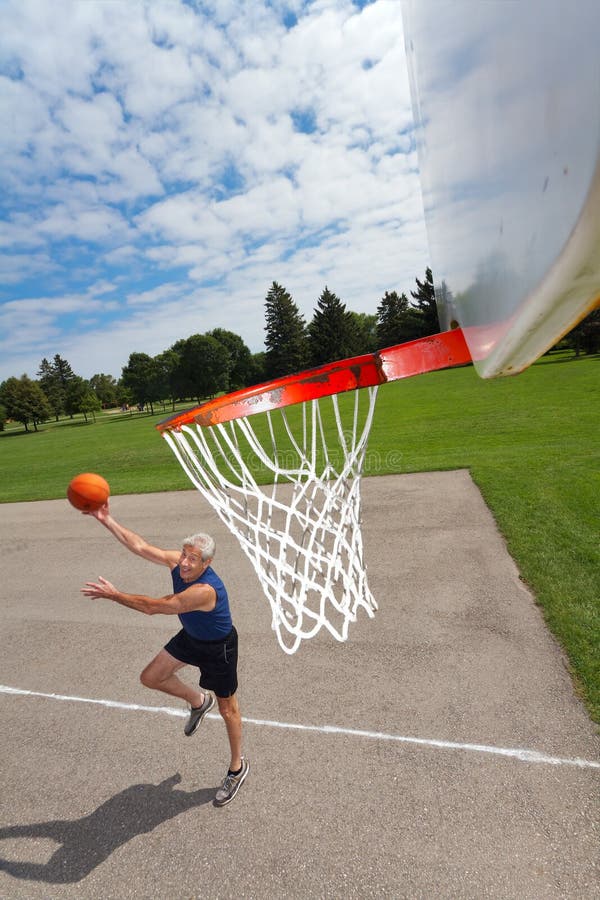 Active senior man shoots hoops on a basketball court outdoors. Background is grass, trees, and blue sky with fluffy clouds. Dynamic shadow. Tilted wide angle view with net large in foreground. Vertical format and copy space. Active senior man shoots hoops on a basketball court outdoors. Background is grass, trees, and blue sky with fluffy clouds. Dynamic shadow. Tilted wide angle view with net large in foreground. Vertical format and copy space.