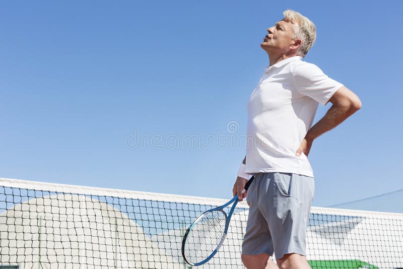 Uomo maturo che fa smorfie con il mal di schiena mentre tenendo la racchetta di tennis contro il chiaro cielo blu il giorno soleg immagine stock libera da diritti Uomo maturo che fa smorfie con il mal di schiena mentre tenendo la racchetta di tennis contro il chiaro cielo blu il giorno soleg immagine stock libera da diritti