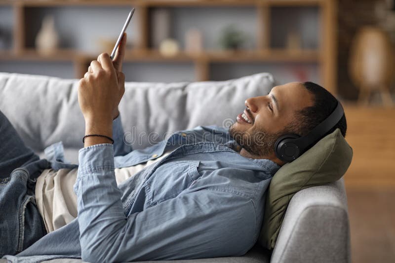 Uomo nero sorridente con cuffie wireless poggiate sulla tavoletta digitale a casa immagini stock
