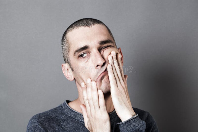 Weird perplexed 30s man acting bored and disturbed, playing with his hands on his face, relaxing himself expressing fatigue and annoyance, grey background indoor. Weird perplexed 30s man acting bored and disturbed, playing with his hands on his face, relaxing himself expressing fatigue and annoyance, grey background indoor