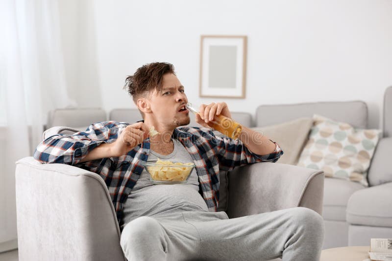 Uomo pigro con la bottiglia di birra e delle patatine fritte che guarda TV immagine stock Uomo pigro con la bottiglia di birra e delle patatine fritte che guarda TV immagine stock