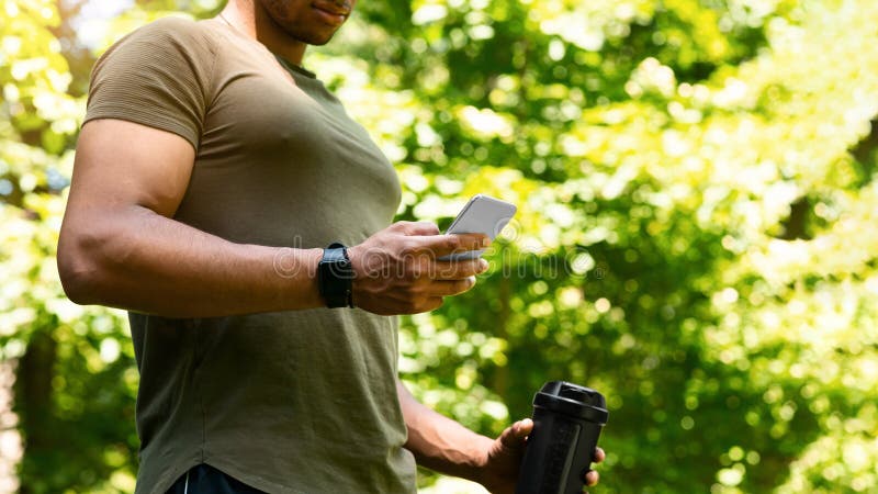 Uomo portante che si prende una pausa dal suo addestramento e naviga su smartphone nello spazio vuoto del parco immagine stock