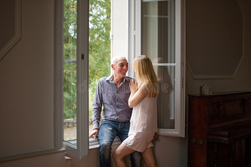 Uomo senior in jeans e camicia che abbracciano la sua giovane moglie bionda che sta vicino alla finestra nella loro casa durante fotografia stock libera da diritti Uomo senior in jeans e camicia che abbracciano la sua giovane moglie bionda che sta vicino alla finestra nella loro casa durante fotografia stock libera da diritti