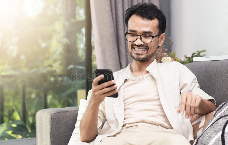 Uomo sorridente e seduto sul divano a casa con l'uso del telefono fotografie stock