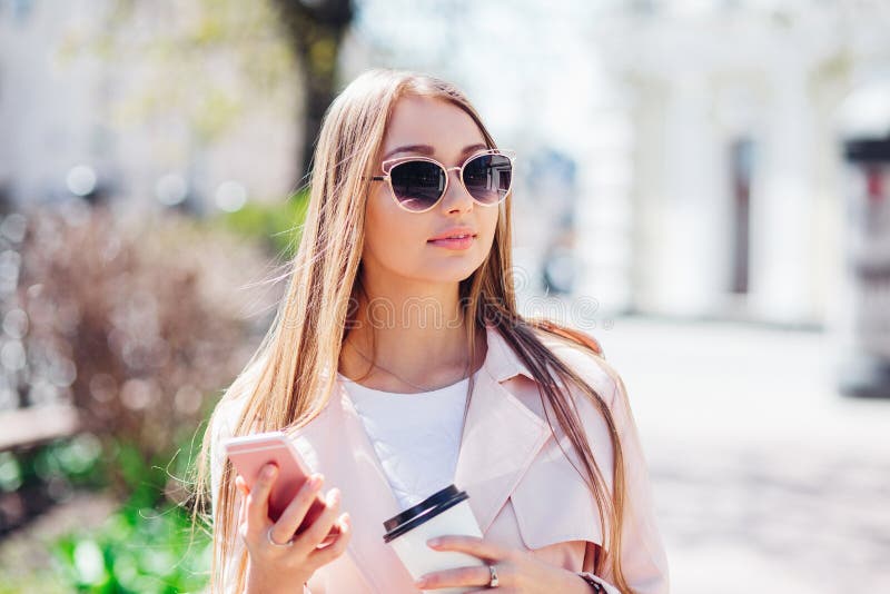 Upper class woman . Fashionable woman texting outdoors. Fashion woman in a sunglasses and pink jacket with coffee.