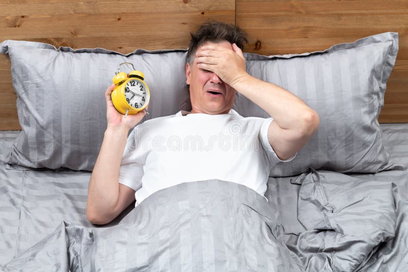 Upprörd man som ligger i sängen och som håller en gul larmklocka i handen och tillverkar facepalm. koncept när jag försovit mig ti royaltyfri fotografi